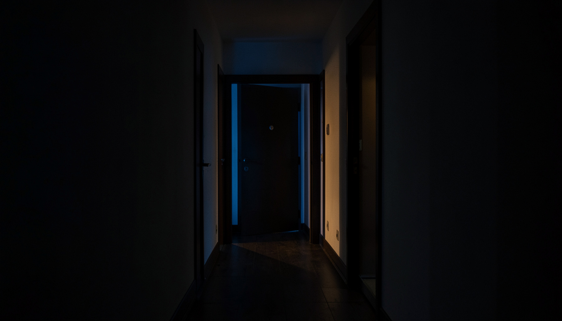 Dark, moody photograph of a quiet Budapest apartment building hallway at dusk, deep shadows and warm amber light spilling from a doorway