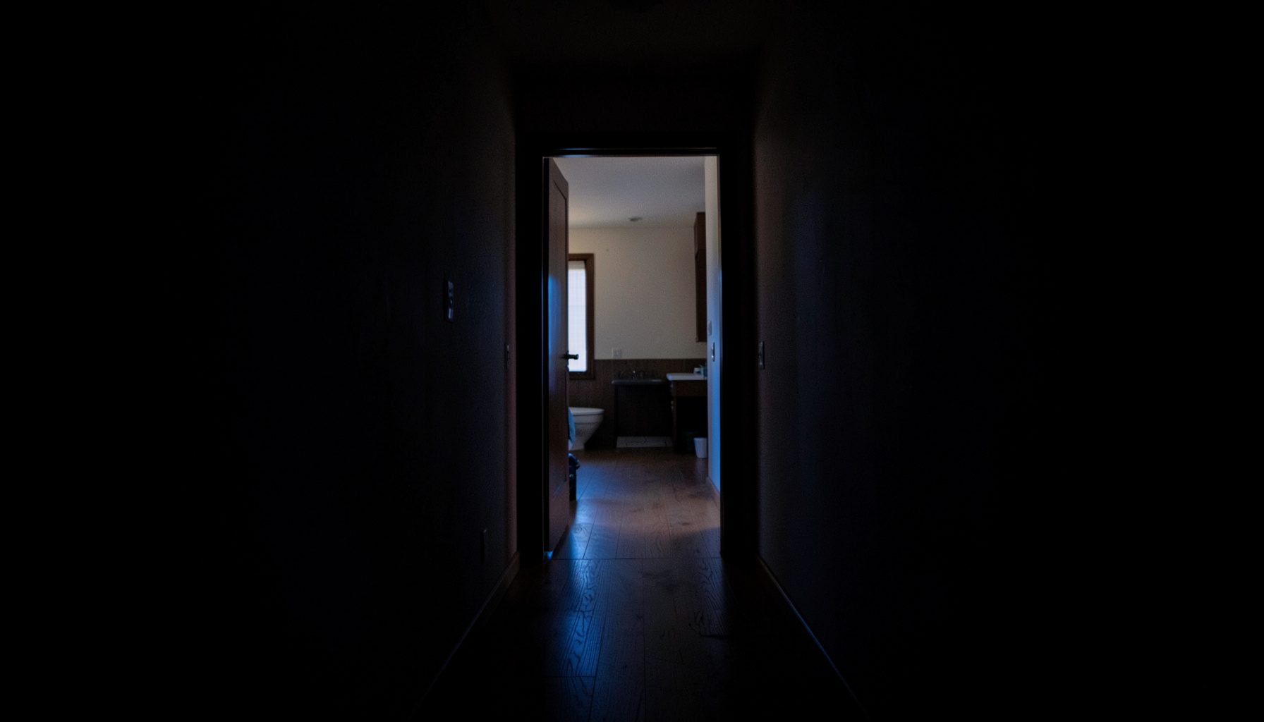 Dark interior hallway with deep shadows, leading toward a softly lit apartment doorway